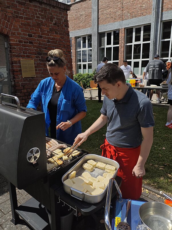Zwei Personen bereiten an einem Grill Würstchen und Grillkäse für das gemeinsame Sommerfest zu.