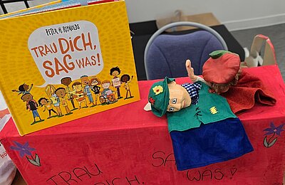 Bilderbuch „Trau dich, sag was!“ steht auf einem Tisch; daneben liegen zwei Handpuppen auf einer roten Box.