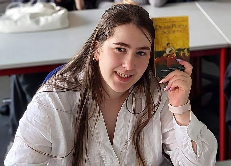 Ein junges Mädchen mit langen Haaren, Ohrringen hat in ihrer linken Hand ein Buch mit dem leicht verschwommenen Titel "DEAD POETS SOCIETY", das sie in Richtung der Kamera hält.
