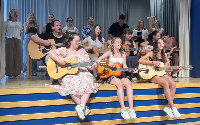 Eine Gruppe von Personen sitzen und stehen auf einer Bühne, spielen Gitarre und singen. Sie geben eine musikalische Darbietung bei einer Abschlussfeier zu geben.