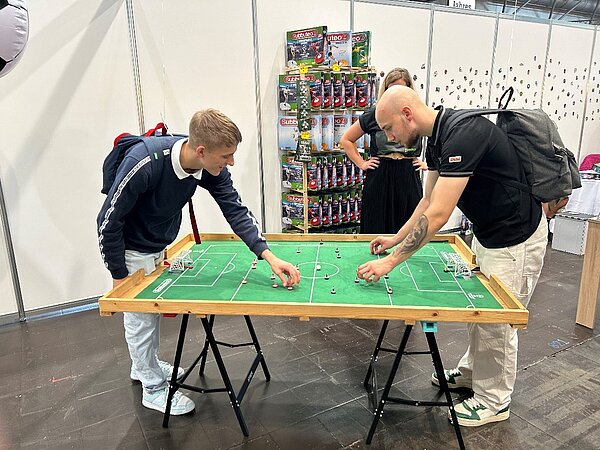 Ein besonderer Tag in Leipzig - Exkursion zur „Hobbymesse“ Zwei Schüler stehen über einen Subbuteo-Tisch (Tischfußball) gebeugt und richten die Figuren aus. Beide jungen Männer haben Rücksäcke auf, der rechte hat auf seinem linken Unterarm ein großes Tattoo eines Totenkopfes. Im Hintergrund steht eine Frau, die dem Spiel zuschaut. Hinter ihr ist ein Regal mit verschiedenen Spielfiguren aufgebaut, die offensichtlich zu verkaufen sind.