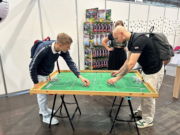 Ein besonderer Tag in Leipzig - Exkursion zur „Hobbymesse“ Zwei Schüler stehen über einen Subbuteo-Tisch (Tischfußball) gebeugt und richten die Figuren aus. Beide jungen Männer haben Rücksäcke auf, der rechte hat auf seinem linken Unterarm ein großes Tattoo eines Totenkopfes. Im Hintergrund steht eine Frau, die dem Spiel zuschaut. Hinter ihr ist ein Regal mit verschiedenen Spielfiguren aufgebaut, die offensichtlich zu verkaufen sind.