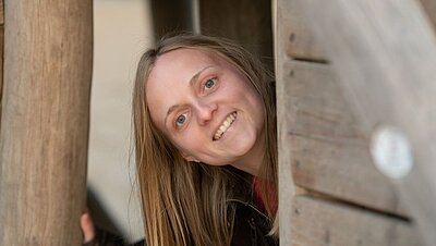 Eine lächelnde Person mit langen, blonden Haaren schaut freundlich aus einer Holzkonstruktion oder Tür heraus.