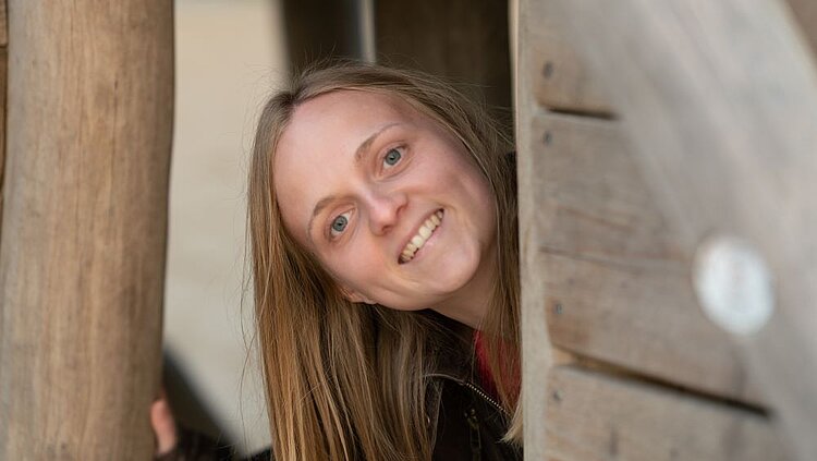 Eine lächelnde Person mit langen, blonden Haaren schaut freundlich aus einer Holzkonstruktion oder Tür heraus.