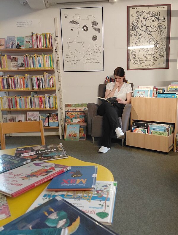 Eine junge Frau sitzt in einem Sessel in einer Bibliothek oder einem Lesebereich und liest ein Buch. Um sie herum stehen Regale mit Kinderbüchern, und an der Wand hängen Bilder.