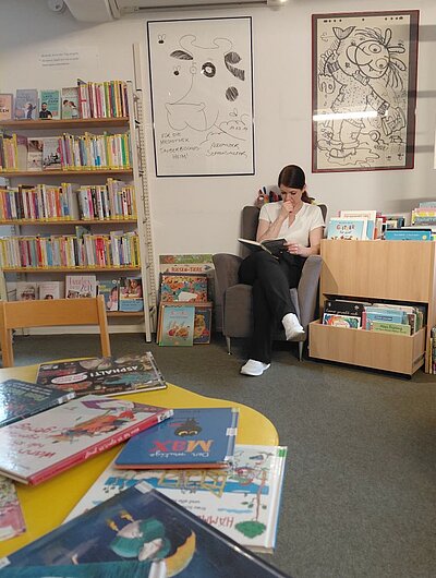 Eine junge Frau sitzt in einem Sessel in einer Bibliothek oder einem Lesebereich und liest ein Buch. Um sie herum stehen Regale mit Kinderbüchern, und an der Wand hängen Bilder.