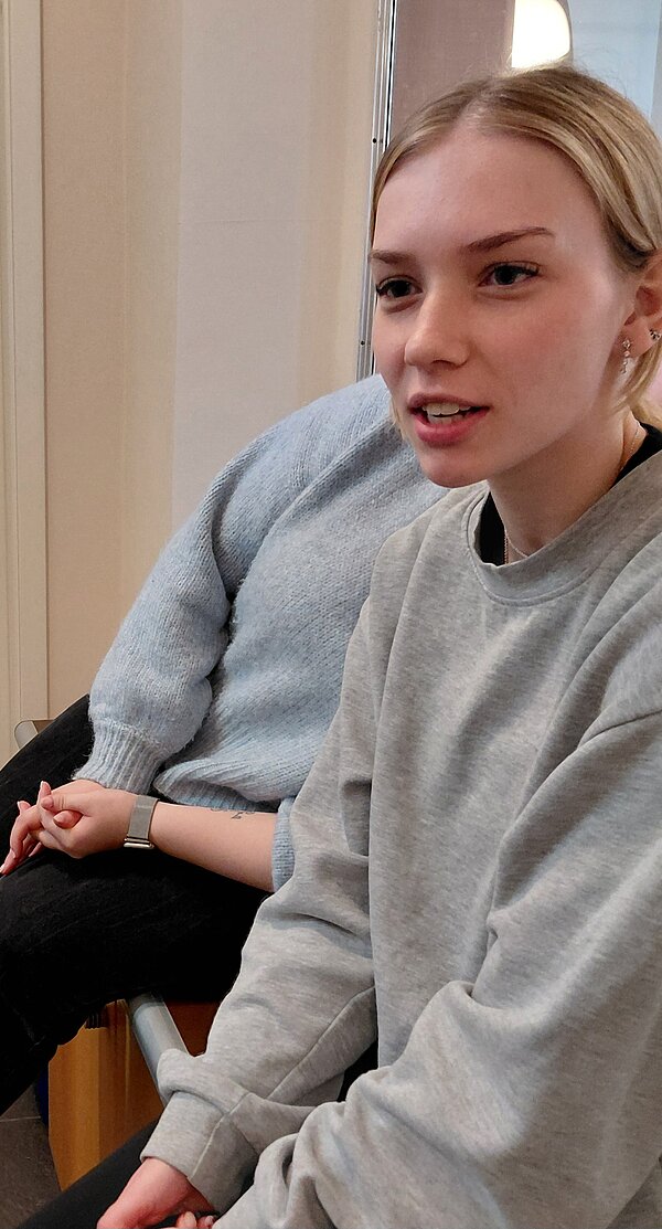 Porträt einer jungen Frau im grauen Sweatshirt, die im Gespräch ist; hinter ihr sitzt eine weitere Person in hellblauem Pullover.