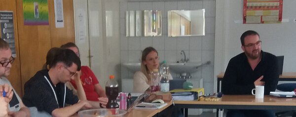 In einem Klassenzimmer sitzen mehrere Schüler an der Seite an Tischen. Am Pult sitzt ein Mann in dunkler Kleidung, der gerade spricht, vor ihm eine Kaffeetasse. Dazwischen an der Ecke sitzt eine Frau, die zuhört.