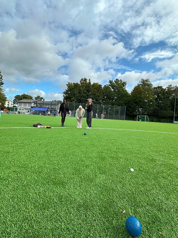 Together in motion – Bewegung, Spaß und Teamgeist Drei Personen spielen auf einem grünen Kunstrasenplatz Boule. Eine Person in der Mitte beugt sich vor, um einen Ball zu werfen, während zwei weitere Personen zuschauen. Im Vordergrund liegen eine kleine weiße und eine blaue Boule-Kugel. Im Hintergrund sind Bäume und ein Zaun zu sehen.