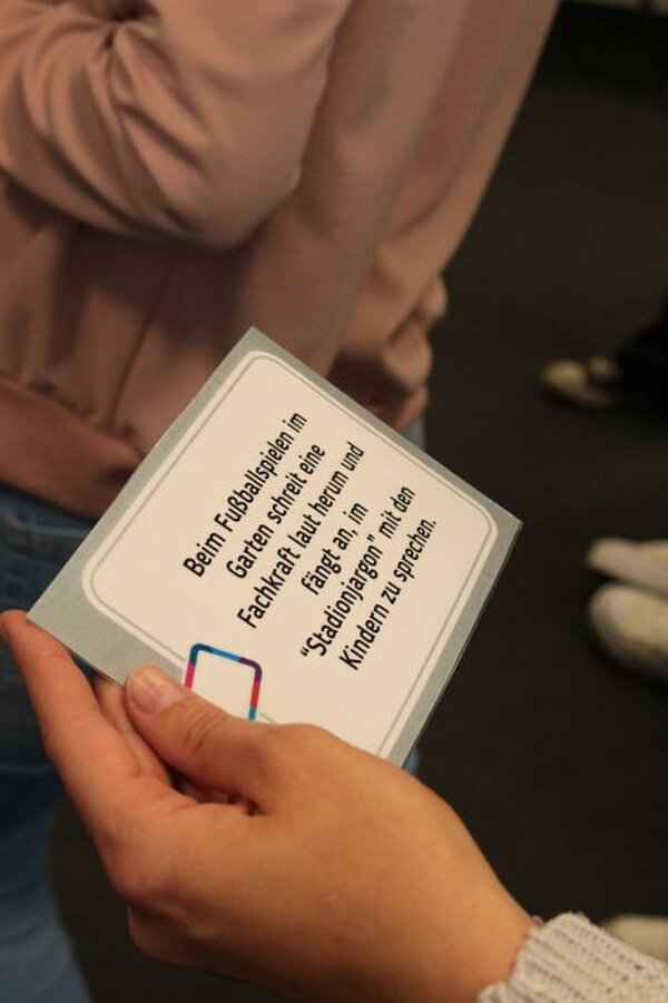Theorie meets Praxis Eine Hand hält eine Karte, auf der steht: "Beim Fußballspielen im Garten schreit eine Fachkraft laut herum und fängt an, im "Stadionjargon" mit den Kindern zu sprechen.