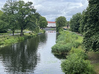 Ein ruhiger Fluss, gesäumt von Bäumen und Sträuchern, führt auf ein großes weißes Gebäude mit rotem Dach zu. Auf dem Wasser sind mehrere bunte Boote zu sehen.