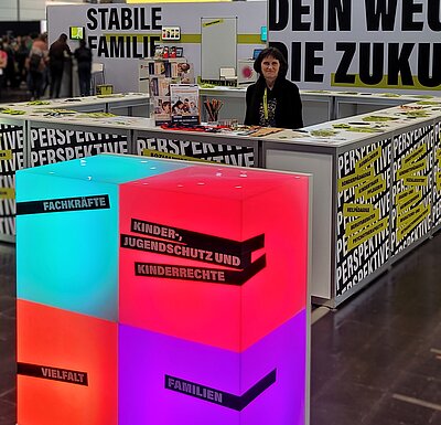 Eine Frau steht hinter dem Tresen eines Messestandes und schaut (etwas kritisch) in die Kamera. Auf der Wand hinter ihr steht in großen Lettern "STABILE FAMILIE". Vor dem Stand sind bunt beleuchtete Quader aufgebaut, auf denen die folgenden Begriffe stehen: FACHKRÄFTE, KINDER-, JUGENDSCHUTZ UND KINDERRECHTE, VIELFALT, FAMILIEN.