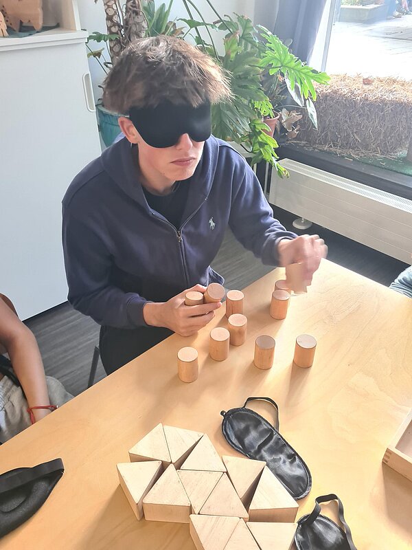 Ein Schüler, der eine Augenbinde trägt, hält mehrere Holzzylinder in der Hand und versucht, diese auf dem Tisch vor ihm zu positionieren. Daneben liegen noch Dreiecke aus Holz sowie weitere Augenbinden.