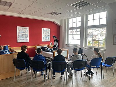 Die Schülerinnen und Schüler sitzen in einem hellen, großen Unterrichtsraum der Euro Akademie Hohenstein im Halbkreis auf blau gepolsterten Stühlen, mit chromfarbenen Beinen vor zwei Pflegebetten. Auf dem linken liegt eine lebensgroße Übungspuppe, auf dem rechten eine Schülerin. An der Wand, die dunkelrot gestrichen ist, hängen zwei medizinische Aushänge. 