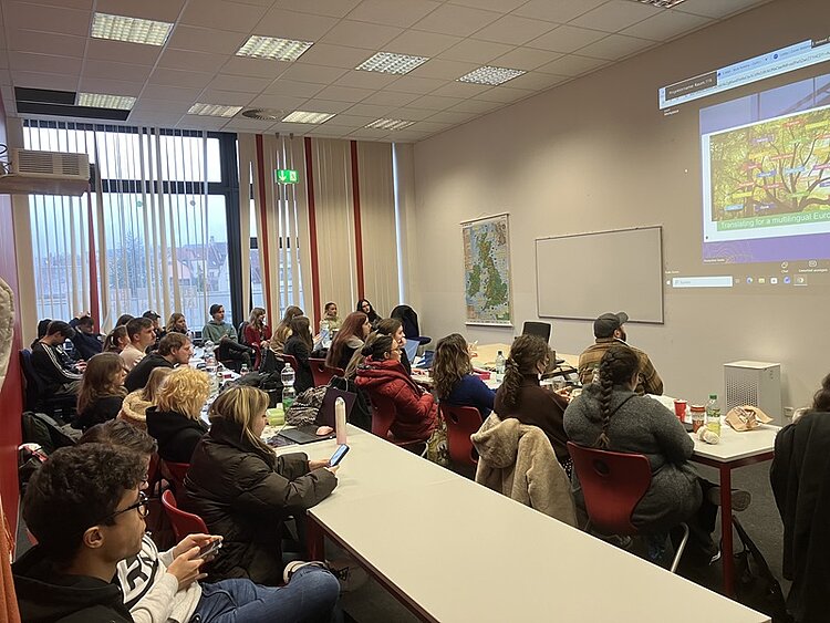 In einem hellen Seminarraum sitzen zahlreiche Auszubildende der Euro Akademie Bamberg an langen Tischen, die in Reihen angeordnet sind. Die Personen sind auf eine Wandprojektion fokussiert, auf der eine bunte Grafik mit Text zu sehen ist. Die meisten Teilnehmenden tragen Winterkleidung wie Jacken und Schals. Im Raum befinden sich große Fenster mit vertikalen Jalousien, durch die Tageslicht einfällt. An der Wand hängt eine Landkarte. Die Atmosphäre wirkt konzentriert und aufmerksam, passend zu einer Online-Veranstaltung der Generaldirektion Übersetzung (DGT) der Europäischen Kommission.