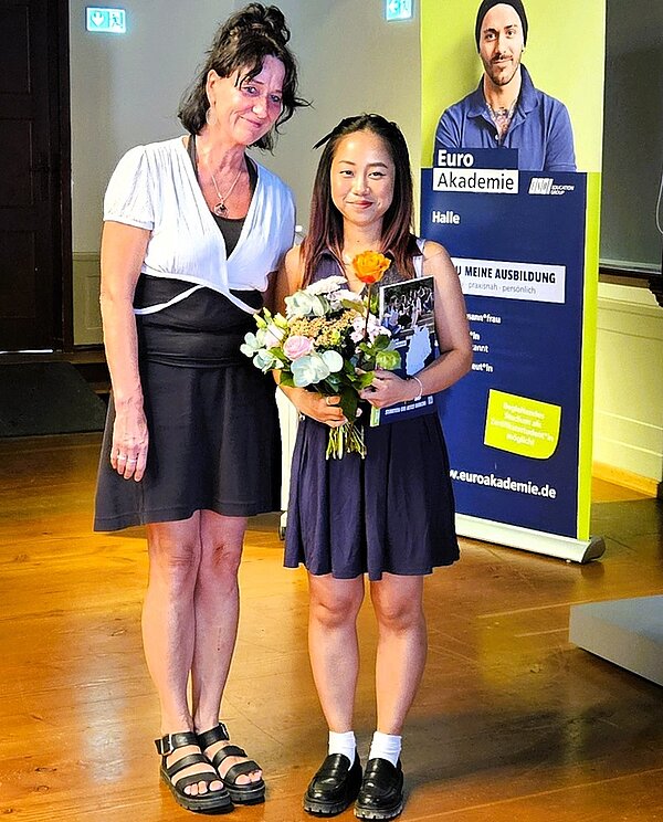 Lehrkraft überreicht einer Absolventin ihr Abschlusszeugnis, einen Blumenstrauß und eine Rose. 