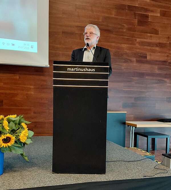 Ein Mann steht an einem Pult mit der Aufschrift "martinushaus" und hält eine Rede. Auf dem Podium stehen Blumenarrangements mit Sonnenblumen. Im Hintergrund ist eine Leinwand mit einer Präsentation zu sehen.