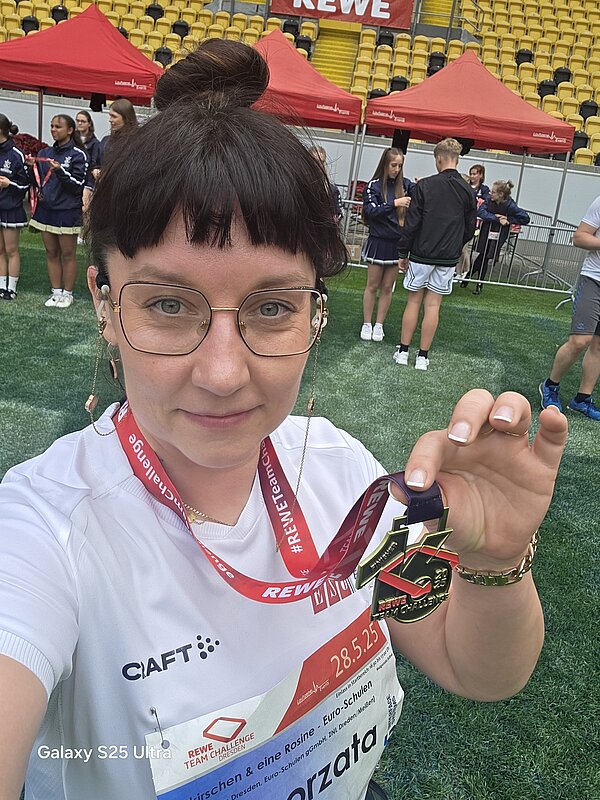 Selfie einer Teilnehmerin, die stolz ihre Finisher-Medaille der REWE Team Challenge zeigt. Im Hintergrund: das Stadion mit roten Zelten und Cheerleadern.