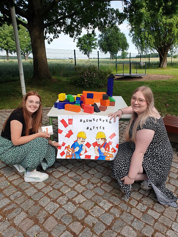 Ein erlebnisreicher Vormittag voller Spiel, Kreativität und Gemeinschaft Zwei junge Frauen knien vor einem Tisch mit bunten Holzklötzen und einem selbstgestalteten Schild mit der Aufschrift "Baumeister Battle" im Grünen.