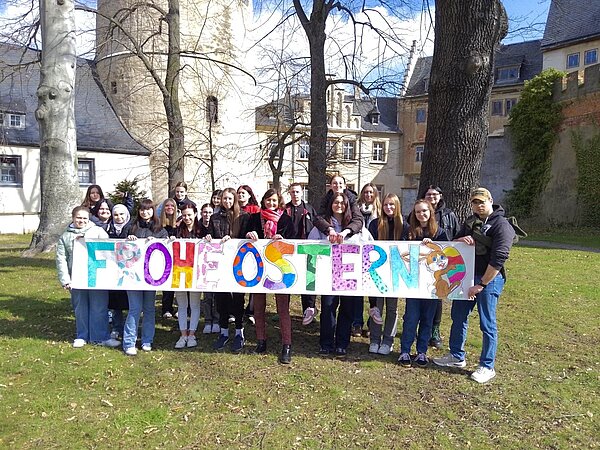 Eine Gruppe von Schülern steht auf einer Wiese vor schlossartigen Gebäuden und hält ein großes Banner mit bunten Buchstaben, auf dem Frohe Ostern steht.