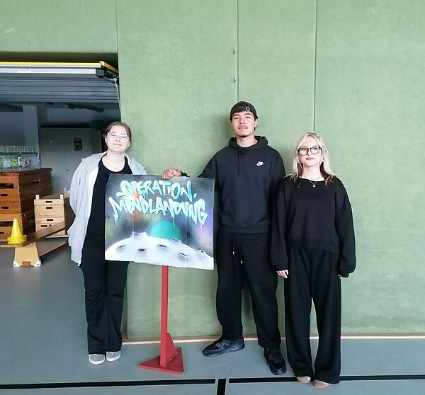 Drei Personen posieren in einer Turnhalle vor einem roten Ständer mit einem UFO-Bild und der Aufschrift ‚Operation: Mondlandung'; im Hintergrund Regale mit Kisten.