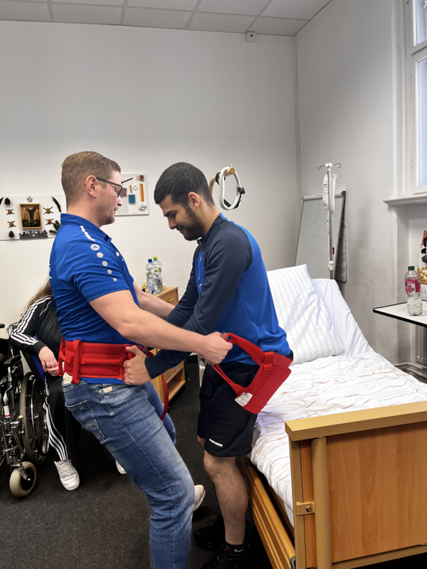 Zwei Personen stehen nebeneinander an einem Pflegebett. Die Person links trägt ein blaues Poloshirt und einen roten Gurt um die Taille, während sie die andere Person mit beiden Händen an einem roten Hebegurt hält. Die zweite Person steht direkt vor dem Bett und trägt ebenfalls ein blaues Oberteil. Im Hintergrund ist eine weitere Person im Rollstuhl zu sehen. Das Zimmer ist mit einem Pflegebett, einem Nachttisch mit Wasserflasche und weiteren Gegenständen sowie einer weißen Wand mit Fenster ausgestattet.