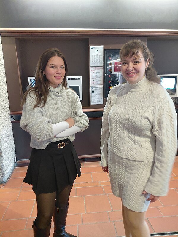 Zwei junge Auszubildende stehen nebeneinander in einem Innenraum der Universitätsbibliothek Würzburg. Die Frau links trägt einen hellgrauen, grob gestrickten Pullover mit hochgekrempelten Ärmeln, einen schwarzen Faltenrock, schwarze Strumpfhosen und schwarze Stiefel. Sie hat lange, braune Haare, die locker über die Schultern fallen. Die Frau rechts trägt ein hellbeiges, grob gestricktes Kleid mit Zopfmuster, das bis knapp über die Knie reicht. Beide Frauen stehen vor einem dunklen Holzrahmen, in dem mehrere Informationsbildschirme und Aushänge sichtbar sind. Der Boden besteht aus rotbraunen Fliesen. Die Szene entstand im Rahmen einer Exkursion der angehenden Erzieher der Euro Akademie Tauberbischofsheim in der Universitätsbibliothek Würzburg.