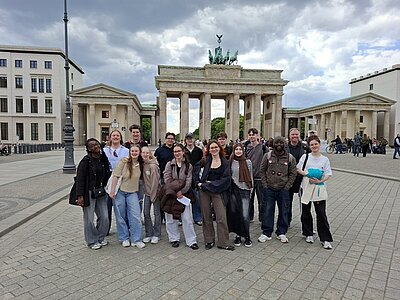Die Gruppe der Azubis Menschen steht auf einem gepflasterten Platz vor dem Brandenburger Tor in Berlin. Die Auszubildenden sind locker nebeneinander aufgereiht und blicken in Richtung Kamera. Im Hintergrund sind das Brandenburger Tor mit der Quadriga sowie zwei große, helle Gebäude zu sehen. Der Himmel ist bewölkt.
