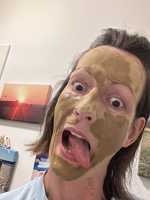 Eine Lehrkraft verzieht das Gesicht zu einer witzigen Grimasse und streckt dabei die Zunge heraus, während die Heilerde auf ihrem Gesicht bereits deutlich angetrocknet ist. 