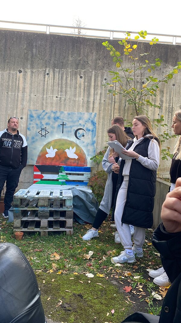 Eine mit einer Israel-Flagge und Palästina-Flagge bemalte Holzpalette steht für einem Holzbrett, auf welchem eine Friedenstaupe und die Symbole des Christentums, des Islams und des Judentums aufgemalt sind. Rundherum stehen Schüler*innen des Projekts.