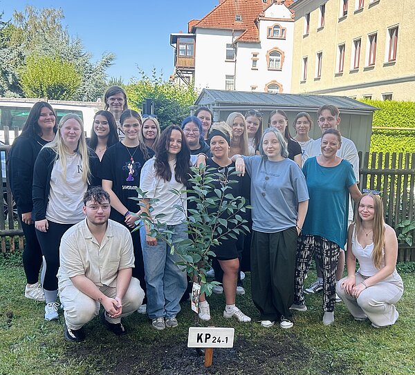 Grüner Schulstart 2025/26 an der Euro Akademie Altenburg Eine Gruppe von Menschen steht im Freien um einen kleinen Baum. Sie posieren lächelnd für ein Gruppenfoto. Der Baum hat ein Schild mit der Aufschrift "KP 24-1". Die Gruppe besteht aus jungen Erwachsenen und sie stehen auf einem Rasen mit einem Gebäude im Hintergrund.