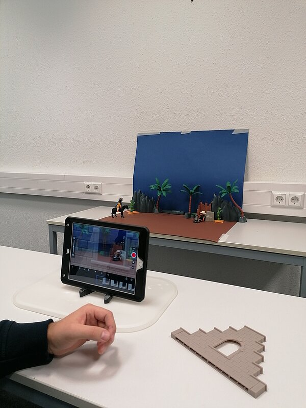 MeKoSax-Projekt „Trickfilmproduktion“ Eine Schülerin filmt eine aus Playmobil gebaute Küste mit Palmen und Felsen, welche auf einem hellbraunen Stück Papier stehen und vor einem hellblauen Hintergrund mit Wolken.
