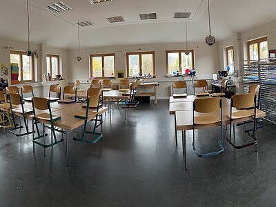 Ein großer, heller Raum mit mehreren Tischen und Stühlen, die in Gruppen angeordnet sind. An den Wänden befinden sich Regale mit verschiedenen Materialien und Dekorationen, darunter ein großes Modell einer Rakete. Der Raum wirkt wie ein Klassen- oder Gemeinschaftsraum, der für verschiedene Aktivitäten genutzt werden kann.