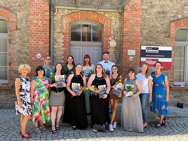 Zeugnisausgabe im Fachbereich Ergotherapie Eine Gruppe von Absolventinnen und Absolventen steht vor einem historischen Backsteingebäude mit einem Schild der Euro Akademie. Sie halten Zertifikate und gelbe Rosen in den Händen. Die Gruppe besteht aus jungen Erwachsenen und Lehrkräften, alle festlich gekleidet. Die Stimmung ist feierlich und fröhlich.
