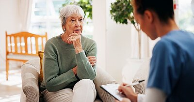 In einem hellen Raum sitzt eine ältere Frau in grünem Pulli und spricht nachdenklich mit einer Person im Vordergrund, die sich Notizen auf einem Klemmbrett macht.