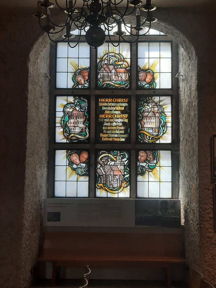 Ethikunterricht mal anders Buntglasfenster mit religiösen Motiven, umrahmt von Engelsköpfen und architektonischen Darstellungen. In der Mitte ein goldener Text, der Christus gewidmet ist. Vor dem Fenster befindet sich eine Holzbank und eine Infotafel.