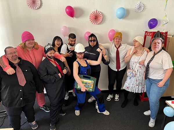 Schüler*innen posieren in humorvollen Kostümen und Verkleidungen in einem mit Luftballons dekorierten Raum.