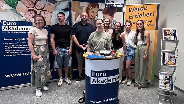 Erfolgreiche Teilnahme am BIT in Wertheim Eine Gruppe von acht Personen steht lächelnd vor einem Informationsstand mit dem Logo der "Euro Akademie". Der Stand hat mehrere Banner, die Informationen über die Akademie und ihre Dienstleistungen enthalten. Auf einem der Banner steht "Werde Erzieher!" und zeigt Bilder von Menschen, die in der Pädagogik arbeiten. Auf dem Tisch vor der Gruppe sind verschiedene Broschüren und Informationsmaterialien ausgelegt. Die Personen scheinen eine positive und einladende Atmosphäre zu vermitteln. Die Website der Akademie, www.euroakademie.de, ist auf den Bannern sichtbar.