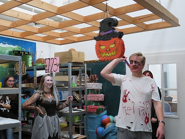 Verkleidete Schülerinnen schauen lachend ihrem verkleideten Lehrer zu, wie er auf eine Kürbis-Piñata schlägt.