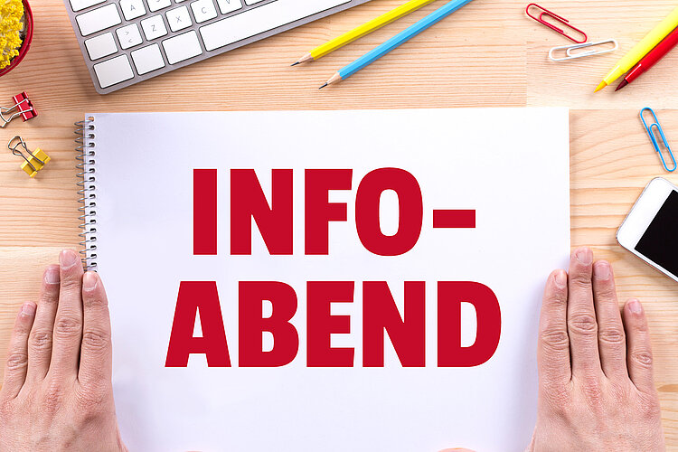 Die Aufschrift "Info-Abend" in roten Lettern steht auf einem Zeichenblock geschrieben, der auf einem Holztisch liegt und neben dem rechts und links die Hände einer Person flach aufliegen. Um den Block verteilt liegen Stifte, Büroklammern und ein Handy und es ist die Ecke einer Tastatur zu sehen.