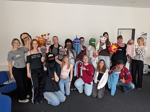 Gruppenfoto mit den Auszubildenden in einem hellen Raum. Sie alle halten ihre eigenen, selbst gestalteten Handpuppen in die Kamera, während sie in die Kamera lächeln.