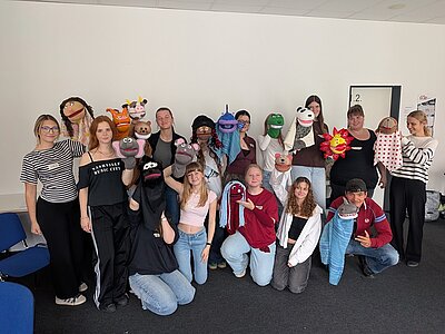 Gruppenfoto mit den Auszubildenden in einem hellen Raum. Sie alle halten ihre eigenen, selbst gestalteten Handpuppen in die Kamera, während sie in die Kamera lächeln.