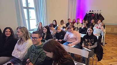 Blick in den großen, hellen Raum, in dem die Schülerinnen und Schüler sitzen und gut gelaunt für ein Bild posieren. 