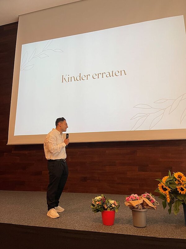 Ein Mann steht auf einer Bühne und spricht in ein Mikrophon. Der Screen hinter ihm trägt den Titel "Kinder erraten". Auf dem Podium stehen Blumenarrangements mit Sonnenblumen.