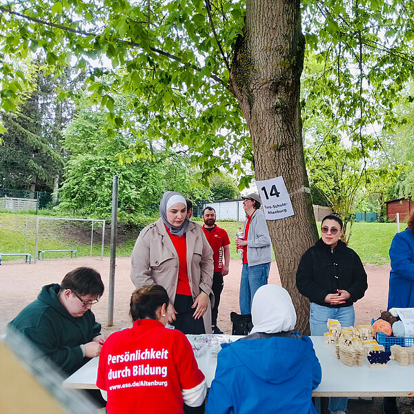Eine Gruppe von Menschen steht im Freien unter einem Baum an einem Tisch mit Bastelmaterialien. Eine Person trägt ein rotes T-Shirt mit der Aufschrift 'Persönlichkeit durch Bildung' und der Website-Adresse www.eso.de/Altenburg. Im Hintergrund sind ein Fußballtor und ein Schild mit der Nummer 14 und der Aufschrift "Euro-Schule Altenburg" zu sehen.