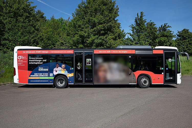 Roter Bus mit Werbung der Euro Akademie Pößneck auf einem Parkplatz vor grünen Bäumen und blauem Himmel.