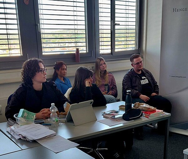 Gruppe von Schüler*innen sitzt an einem Tisch am Fenster und hört einer Präsentation zu.