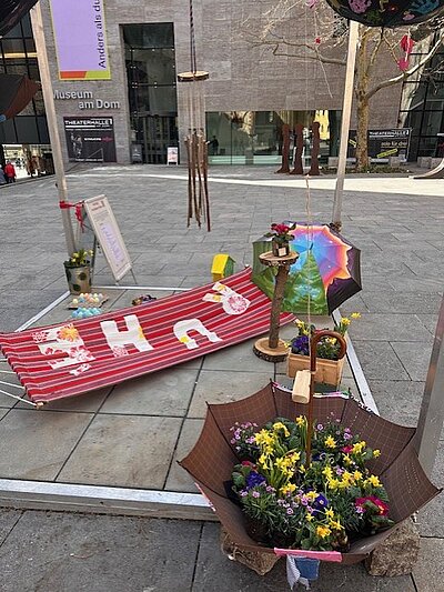 Auf einem Platz vor dem Dom ist Kunst ausgestellt: darunter ein umgedrehter Regenschirm mit Blumen darin; ein bunt bemalter Regenschirm und eine Hängematte mit dem Wort "Ruhe".
