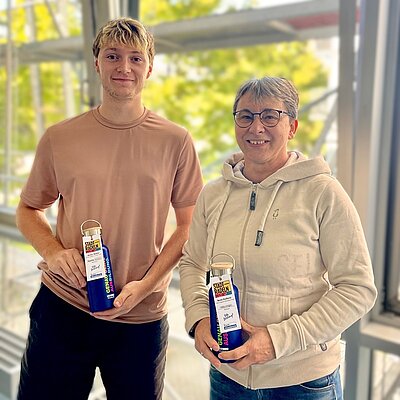 Der Schüler Sascha und die Mitarbeitende Frau Unger halten ihre Auszeichnung, in Form von Thermoskannen, als beste Radlerin und Radler in Händen und posieren für ein gemeinsames Foto. 