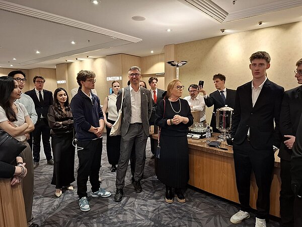 Eine Gruppe von Menschen steht in einem Konferenzraum mit Teppichboden und beigen Wänden. Die Personen tragen überwiegend formelle Kleidung wie Anzüge, Hemden und Kleider. Im Hintergrund befinden sich Tische mit technischen Geräten, darunter ein zylindrisches Metallobjekt mit Kabeln und ein weiteres Gerät mit Glaszylinder. Die Beleuchtung ist hell und gleichmäßig. Einige Personen halten Smartphones in der Hand.